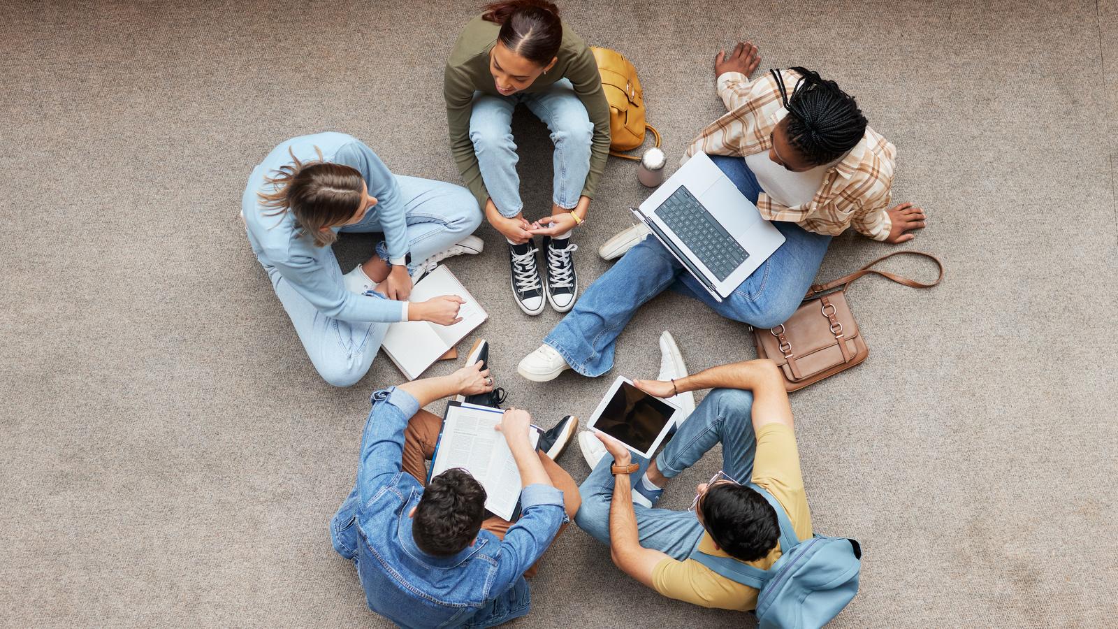 Five people sitting in a circle on the floor, planning together with notebooks, a laptop and a tablet