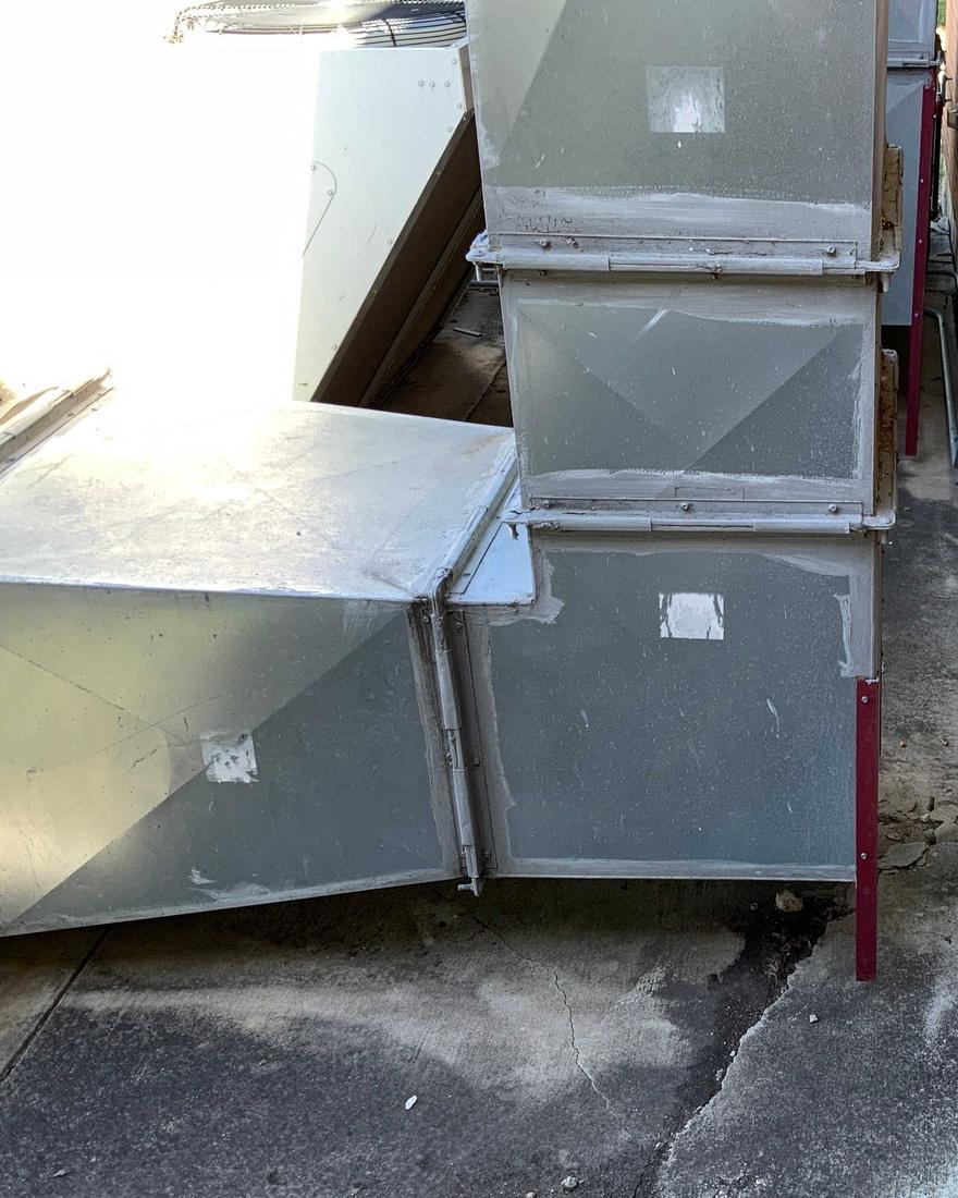 Sections of sheet-metal air duct stacked outside a brick school wall behind a chain-link fence, their seams patched, on a cracked and stained concrete pad