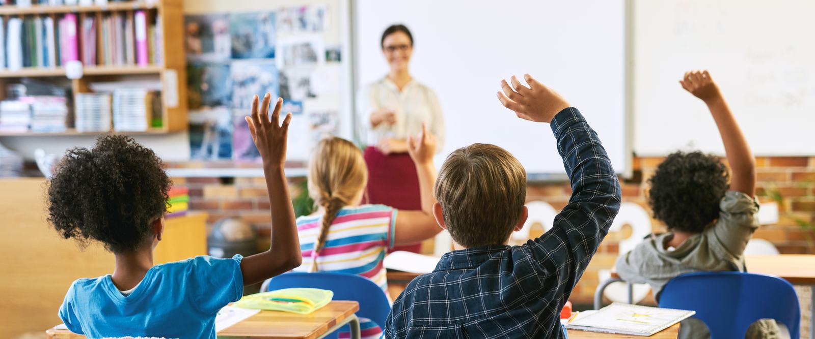 Elementary students raising their hands in a classroom while a teacher stands at the front