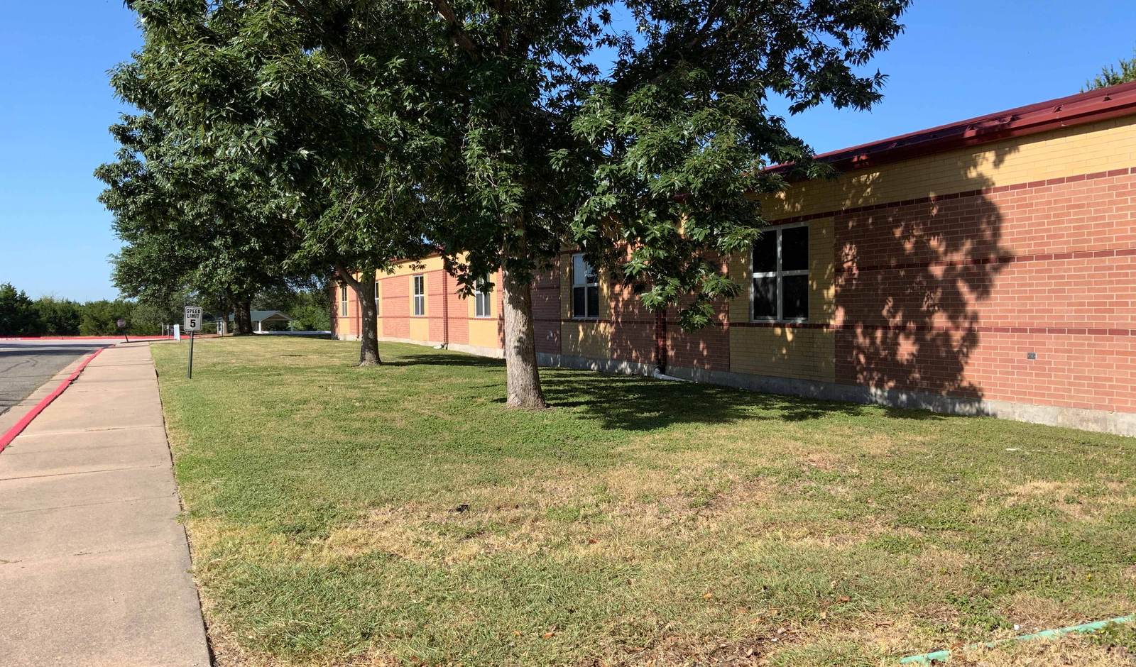 A Manor ISD elementary school on a clear morning, with shade trees along the front lawn and a sidewalk leading to the building