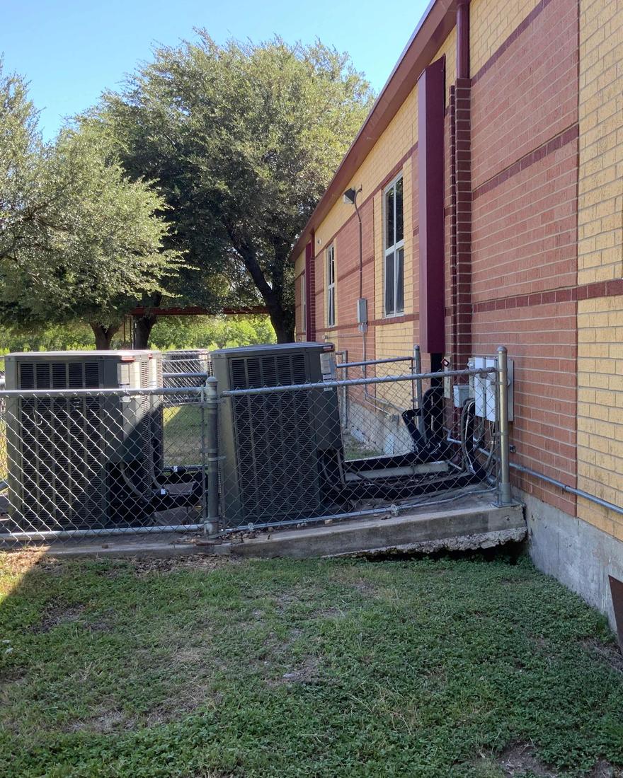 Two ground-level air-conditioning condenser units inside a chain-link pen beside a brick school wall, with electrical conduit strapped to the outside of the building