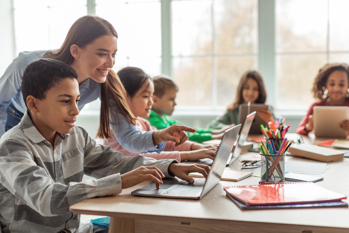 A teacher leaning over a student's shoulder to help with a laptop in a bright classroom, other students working behind them