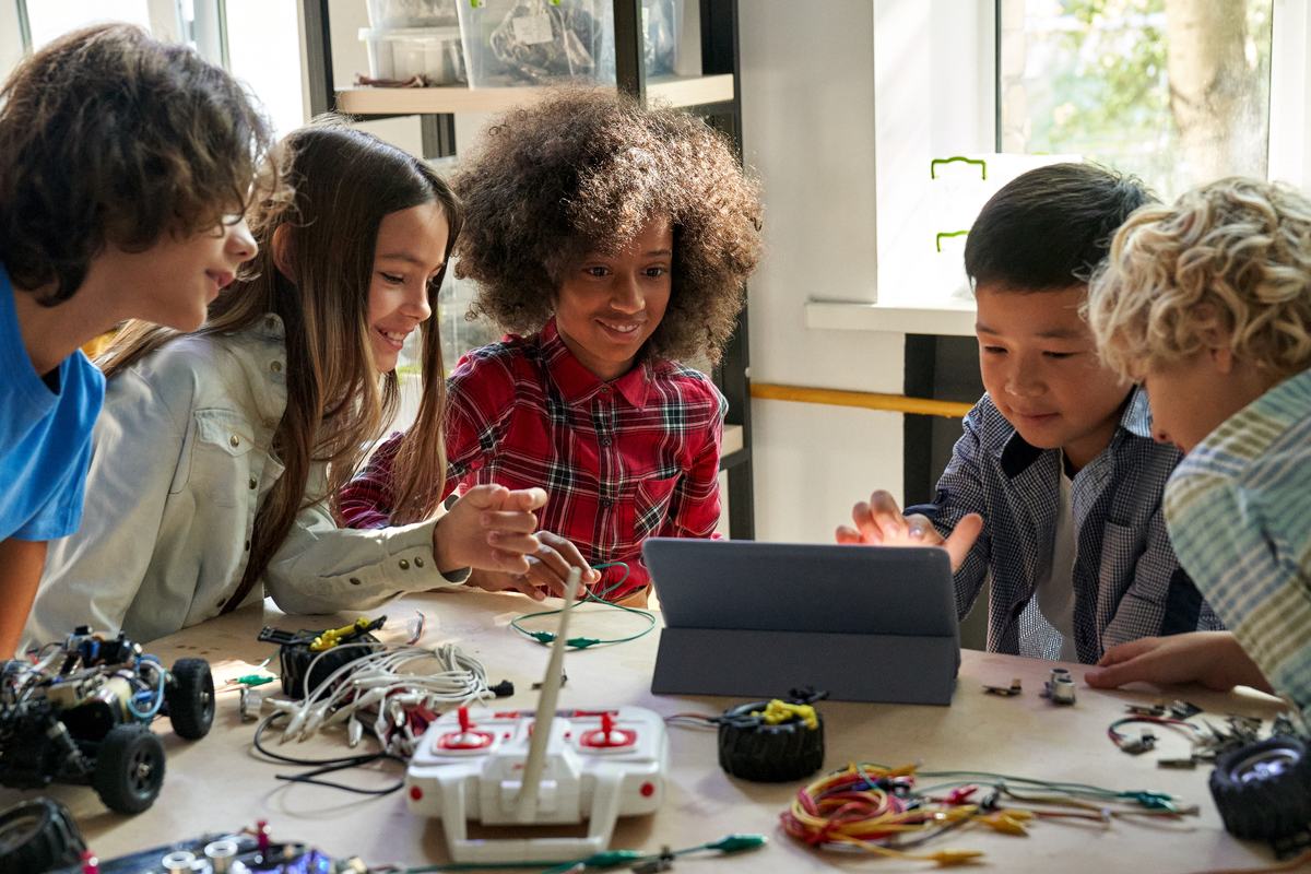 Five elementary students gathered around a tablet at a table covered with robotics parts and wiring