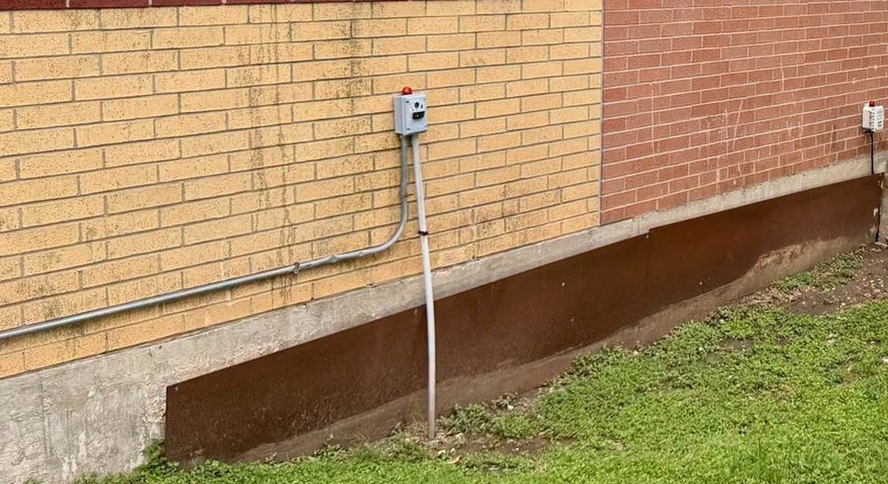 The base of a brick school wall with a stained concrete skirt, electrical conduit and control boxes mounted on the outside