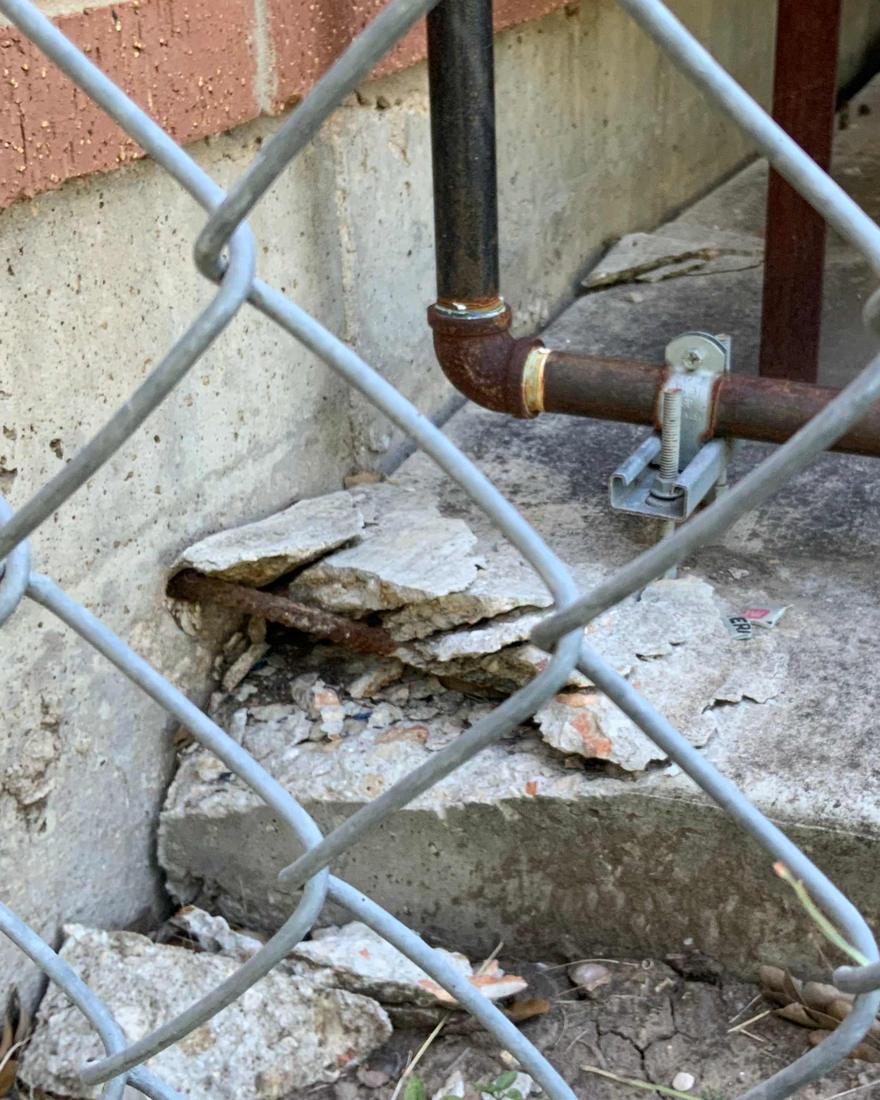 The edge of a concrete foundation slab breaking apart beside a chain-link fence, with rusted steel reinforcing bar exposed and a rusted pipe running along the wall